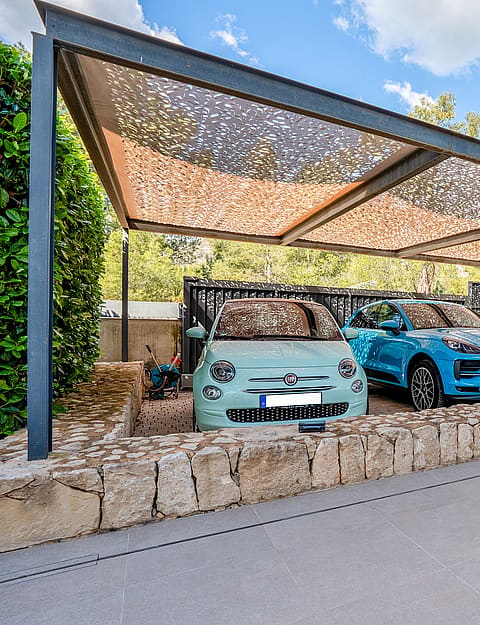 Covered carport with steel frame and patterned shade canopy beside a natural-stone wall.