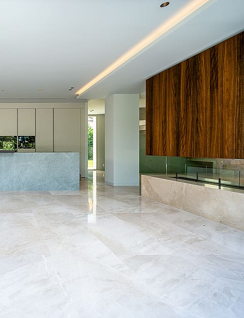 Wide shot of a modern kitchen with a blue-grey stone island, polished light-colored marble flooring, and a linear fireplace.