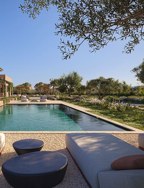 A wide exterior view of a finca with a rectangular swimming pool, gravel patio, sun loungers, and pergola overlooking mountains.