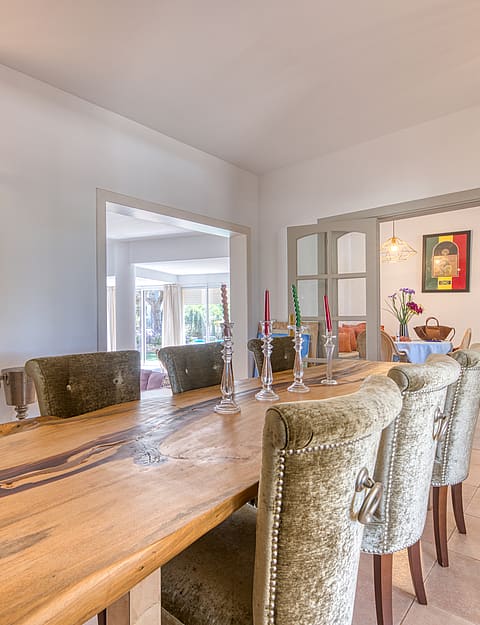 A formal dining room featuring a long live-edge wooden table, upholstered high-back chairs, and neutral stone tile floors.