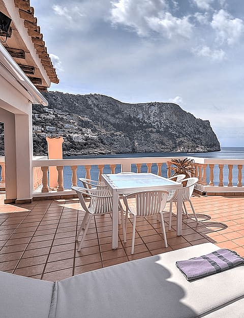 A wide outdoor terrace in Cala Llamp with terracotta floor tiles, a stone balustrade, and white outdoor dining furniture overlooking the sea.