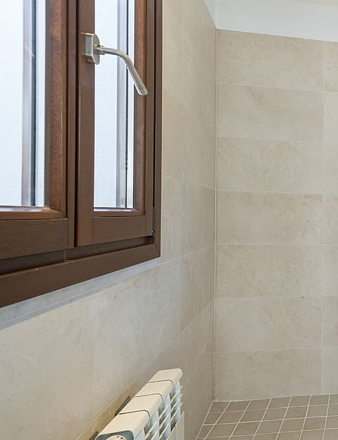 Contemporary bathroom with beige tiling, glass walk-in shower, rainfall head, and a wooden Mallorcan-style window.