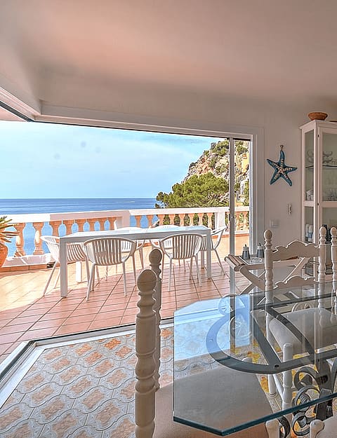 Interior dining room with a glass-top table, white wooden chairs, and a rattan pendant light, opening to a terrace with sea views.