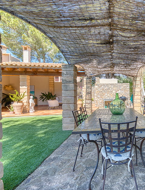 A stone-columned patio with a trellis roof and iron dining set, overlooking a green garden and building exterior.