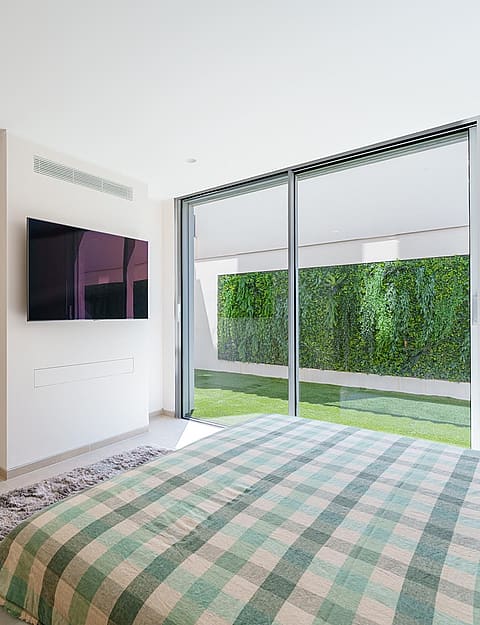 Contemporary bedroom with checkered bedspread, wall-mounted television, and sliding glass doors opening to a green wall.