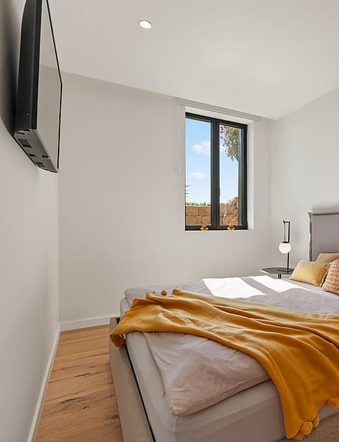 A modern bedroom with an upholstered grey bed, mustard yellow throw blanket, wooden flooring, and a single vertical window.