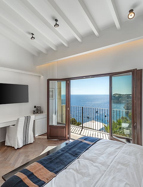 Master bedroom featuring white beamed ceilings, herringbone wood floors, and a balcony opening to Mediterranean sea views.