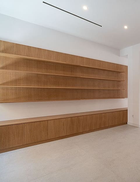 A bright, minimalist open-plan living room with light-colored stone floors, white walls, and long, wall-mounted oak shelving units.