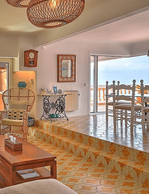 Sunlit living area with tiled floors, rattan armchairs, and a view of Cala Llamp through glass sliding doors.