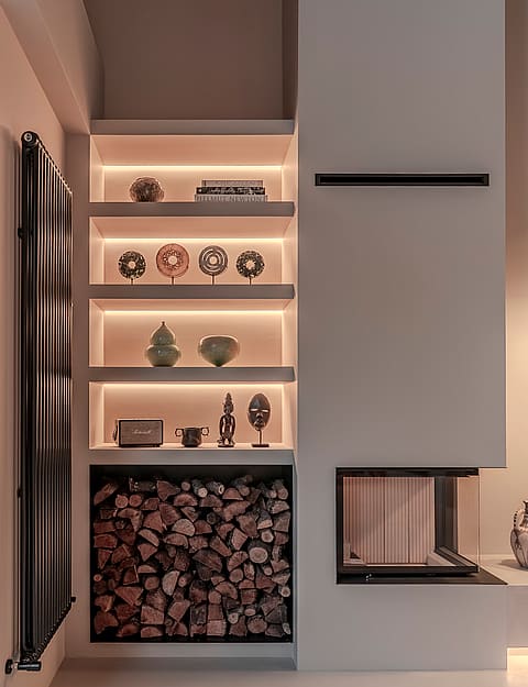 A modern white fireplace with a glass corner unit, recessed backlit shelves displaying artifacts, and a log storage niche.