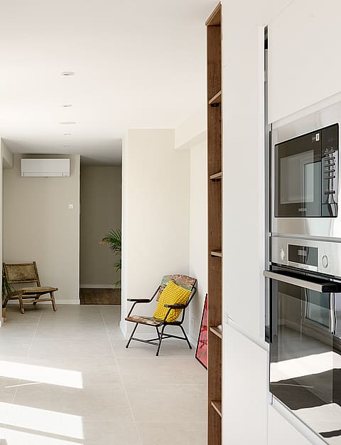 Interior view from a kitchen looking towards a living area with white walls, tiled floors, a stool, and a yellow chair.
