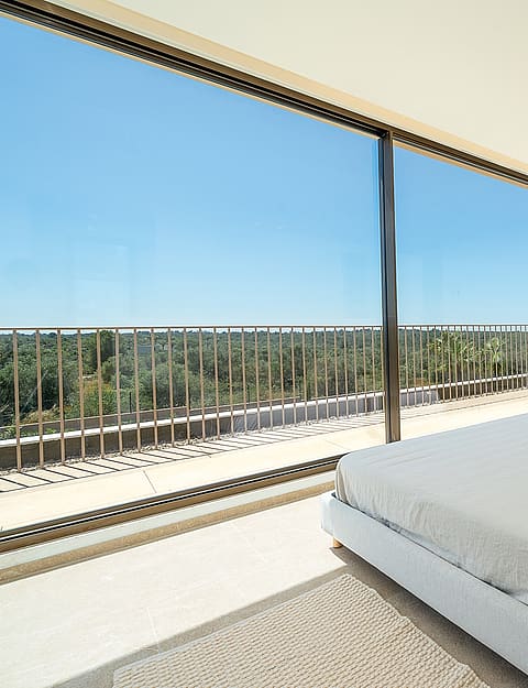 A minimalist bedroom featuring a platform bed with beige bedding, overlooking a sunlit balcony with mountain views.