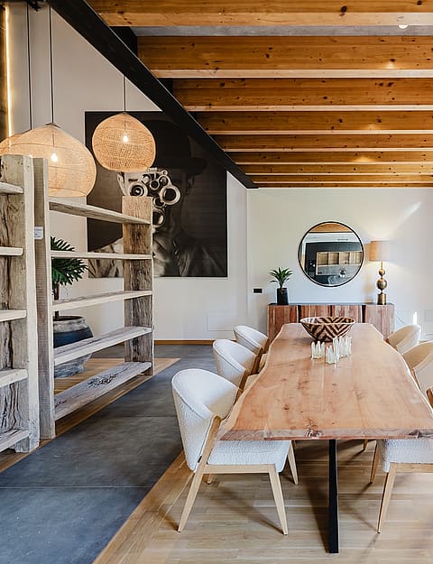 Dining area featuring a long live-edge wooden table, white upholstered chairs, and exposed wooden ceiling beams in a spacious room.