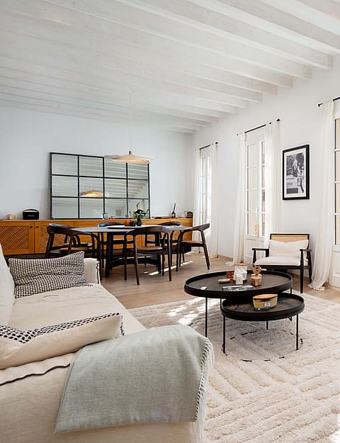 A bright, open-plan living room with white walls, exposed white ceiling beams, a textured area rug, and three sets of French doors leading to a balcony.