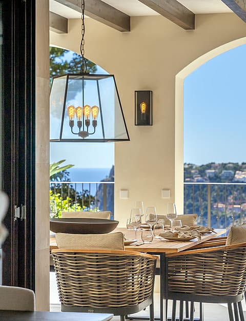 A dining table set with glassware on a covered terrace featuring wicker chairs, wood ceiling beams, and arched sea views.