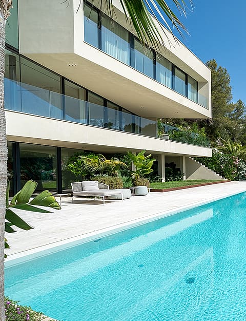 Wide exterior view of a white, multi-level modern villa featuring a long rectangular swimming pool, terrace, and trees.