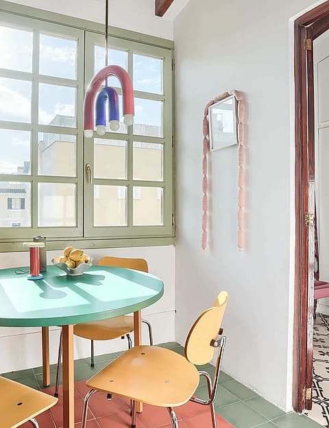 A small dining area with a round green table, wooden chairs, and a window overlooking city buildings in Santa Catalina.