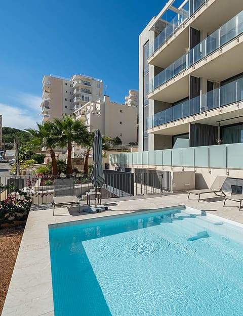 A rectangular outdoor swimming pool with light blue water sits on a stone terrace in front of a modern multi-story apartment building with glass balcony railings.