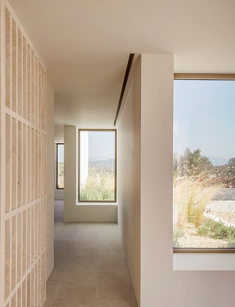 A sunlit hallway featuring light stone flooring, smooth plaster walls, and a vertical timber slatted screen beside large windows.