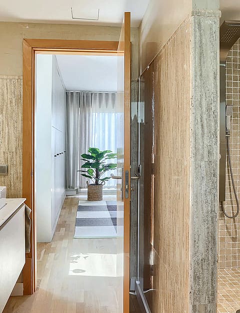 Modern bathroom featuring dual square sinks, stone-patterned tiled walls, and a glass-enclosed shower, leading into a bedroom.
