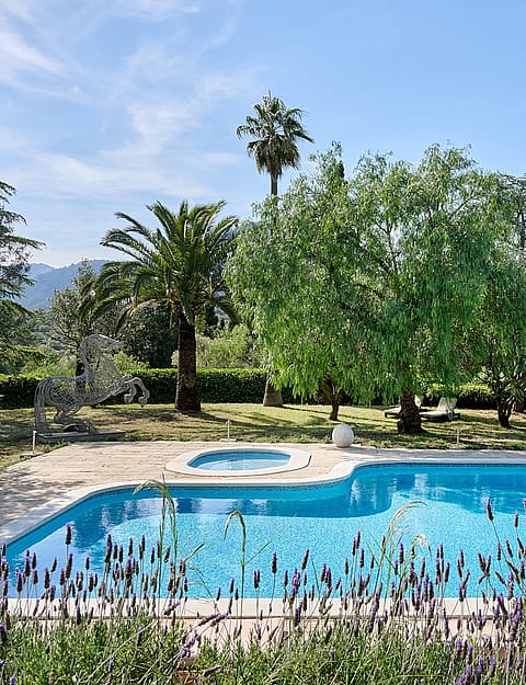 A blue swimming pool sits in a mature garden with palm trees, lavender in the foreground, and mountains in the distance.