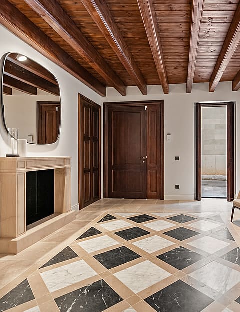 Grand entrance hall featuring dark wood ceiling beams, a checkered marble floor, and a stone fireplace with an organic mirror.