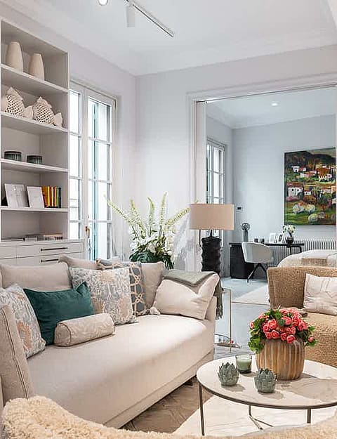 A bright living room featuring a wall-to-wall white shelving unit, a cream sofa with decorative pillows, and an open doorway leading to a secondary room.