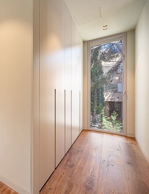 A minimalist dressing room corridor in a Santa Ponsa villa, featuring light oak wood floors, floor-to-ceiling white built-in wardrobes, and expansive glass windows overlooking greenery.