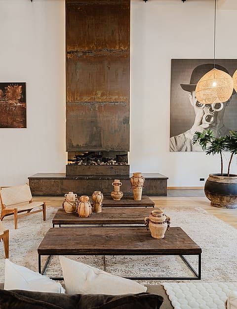 Spacious living room featuring a floor-to-ceiling oxidized metal fireplace, wooden coffee tables, low-profile seating, and woven pendant lights.