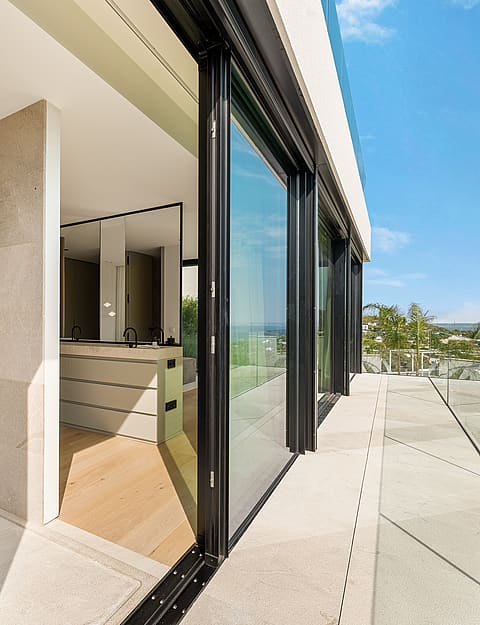 A wide shot looking from a stone-paved terrace into an interior room through sliding glass doors, with a clear blue sky above.