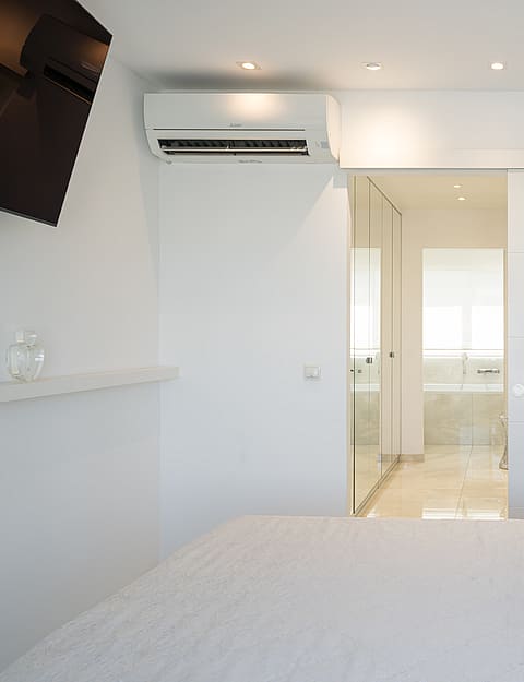 A modern bedroom with a neutral palette, a large bed with a padded headboard, and a sliding door leading to an en-suite bathroom.