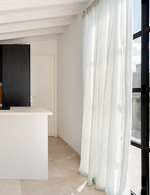 Modern kitchen with a white central island, dark cabinetry, and exposed white ceiling beams, adjacent to a staircase.