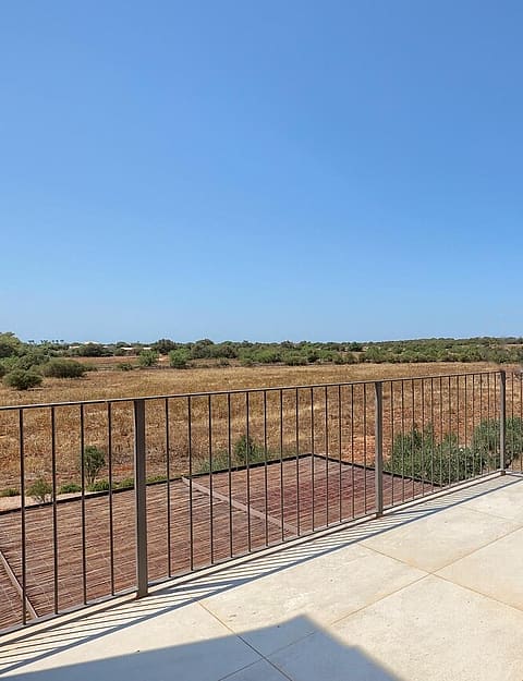 A stone-paved terrace with a metal railing overlooking a dry Mediterranean landscape, distant hills, and a swimming pool below.