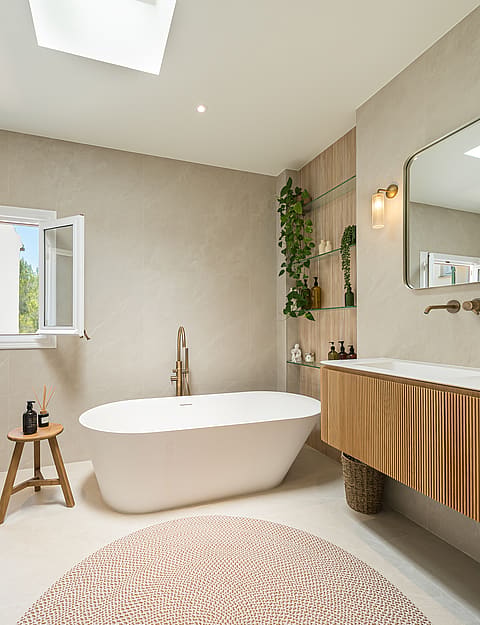 Contemporary bathroom with a freestanding oval bathtub, fluted wood vanity, brass fixtures, and natural light from a skylight.