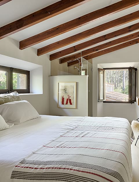 Master bedroom with vaulted ceiling, exposed wooden beams, white bedding, and a decorative chair in an Alaro finca.