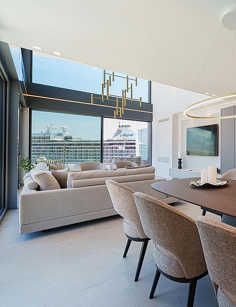 Comedor con mesa ovalada de madera, sillas tapizadas y lámpara circular moderna, abierto hacia el mar.
