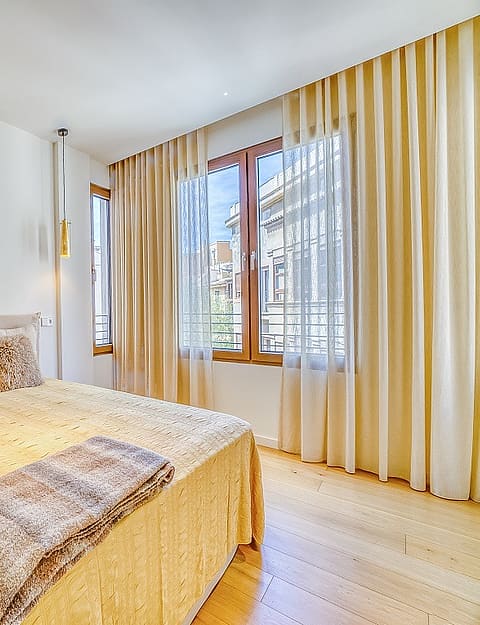 A double bedroom featuring wooden floors, beige curtains, and two hanging pendant lights beside the bed headboard.