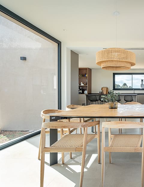Dining room with a wooden table, woven chairs, and a large pendant light, framed by glass sliding doors and stone flooring.
