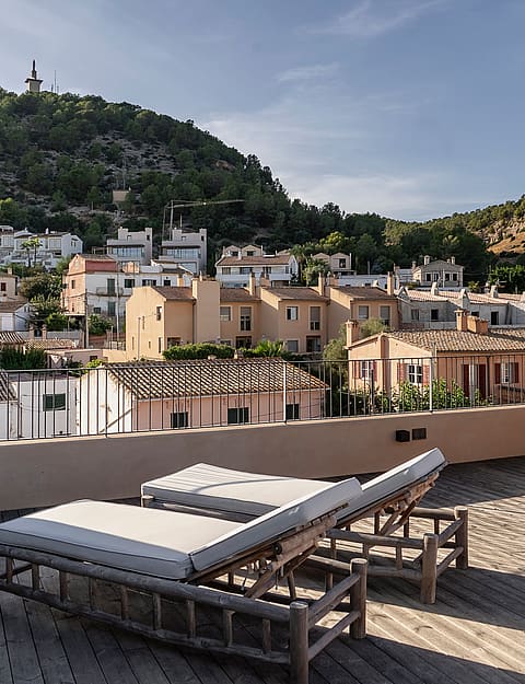 Rooftop terrace featuring two wooden sun loungers with white cushions, overlooking a hillside town in Genova under clear skies.