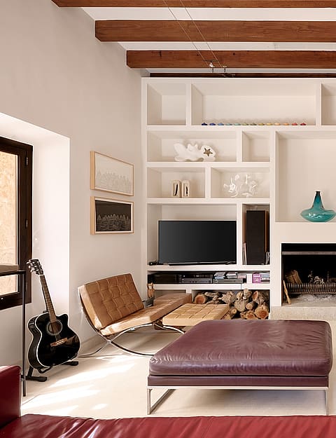 A living area with white walls, wooden ceiling beams, and a floor-to-ceiling white shelving unit built around a fireplace.