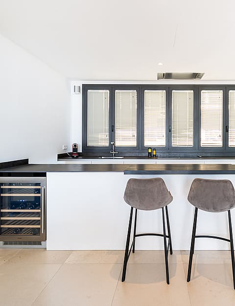 Modern kitchen featuring a white island with two stools, dark countertops, and a built-in wine fridge against a bright window.
