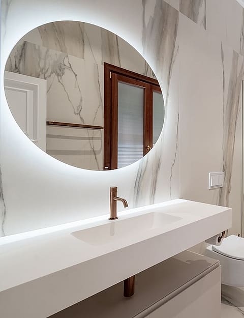 A contemporary bathroom featuring floor-to-ceiling white marble with grey veining, a floating vanity, and a circular LED-lit mirror.