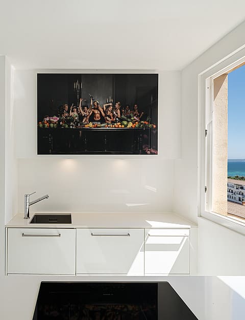 Modern white kitchen featuring a countertop, sink, and high-contrast artwork, positioned next to a large window overlooking the city.