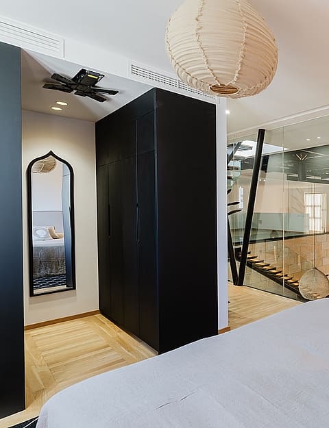 Modern master bedroom featuring a large bed, dark matte wardrobe, and glass walls separating the space from an internal staircase.