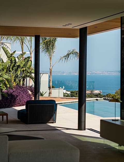 An interior living space looking out toward a swimming pool and the Mediterranean sea through large glass walls.