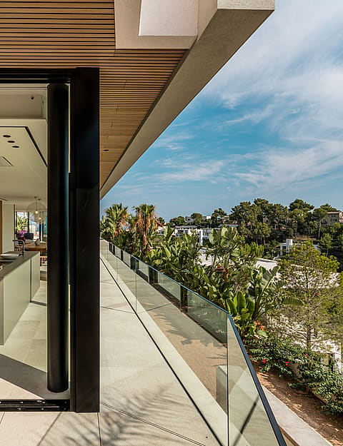 Wide angle view from a modern kitchen looking out onto a long stone terrace with a frameless glass balustrade and greenery.