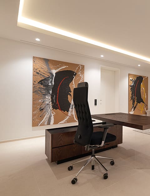 Interior view of an open-plan office space featuring a dark wood desk, contemporary paintings on white walls, and a recessed lounge corner with a grey sofa and gym equipment.