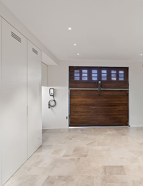 Modern garage featuring polished stone floors, white minimalist cabinets, a dark wood door, and a wall-mounted EV charger.