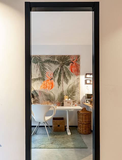 A home office viewed through a black-framed doorway, featuring a botanical mural wall, white desk, and wicker basket.