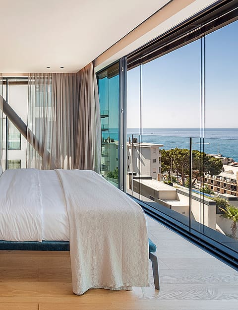 Modern master bedroom with a teal upholstered bed, light oak flooring, and expansive glass walls overlooking the ocean.